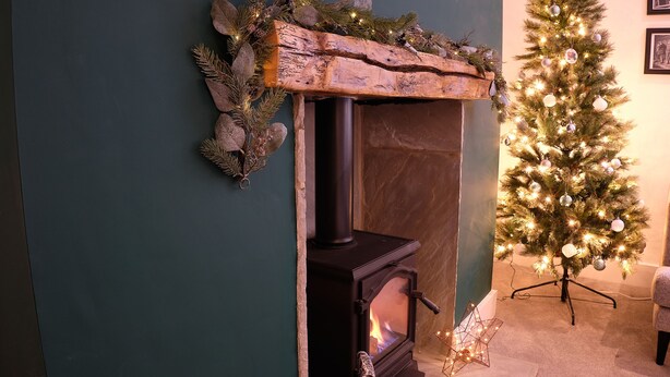 A fireplace with a rustic mantel, a lit fire, and a Christmas tree decorated with lights and ornaments. - Flat Cap Cottage (Skipton)