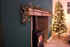 A fireplace with a rustic mantel, a lit fire, and a Christmas tree decorated with lights and ornaments. - Flat Cap Cottage (Skipton)