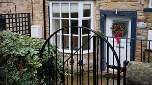A stone building with a black iron gate, a window with a white frame, and a door with a wreath. - Flat Cap Cottage (Skipton)