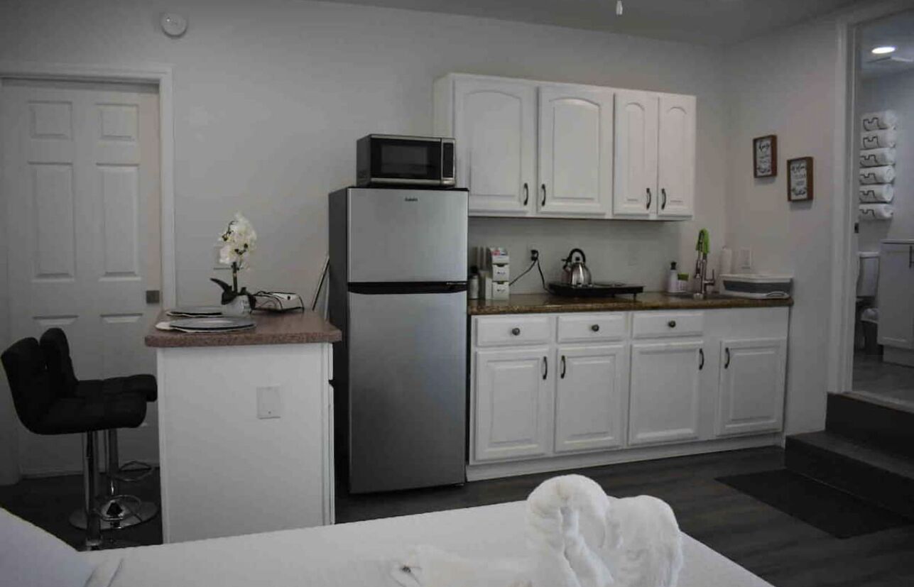 A compact kitchen with white cabinets, a stainless steel refrigerator, a microwave, and a small bar stool.