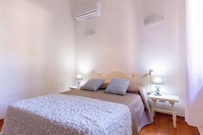 A bedroom with a bed, two bedside tables with lamps, a white blanket, and an air conditioning unit on the wall.