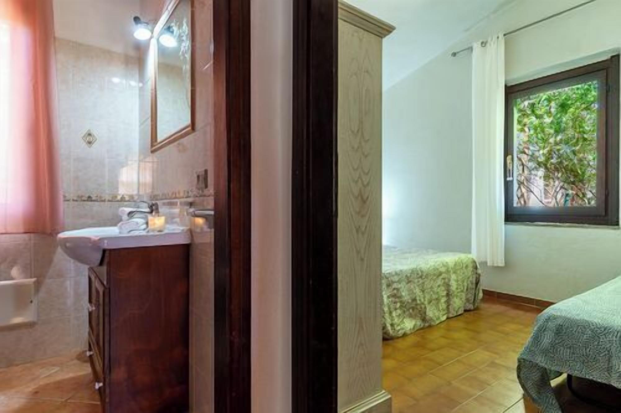 A bathroom with a sink, mirror, and shower. There's a bed with a green bedspread and a window with a view of greenery.
