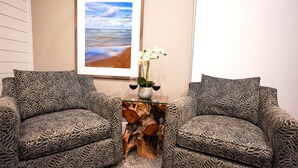 Un área de descanso con cojines estampados, una mesa de vidrio sobre base de madera y un cuadro enmarcado con una escena de playa.