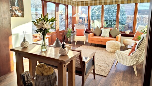 A cozy living room with a wooden table, wicker chairs, a sofa, and large windows offering a view of greenery. - Yewtree House (Penrith)