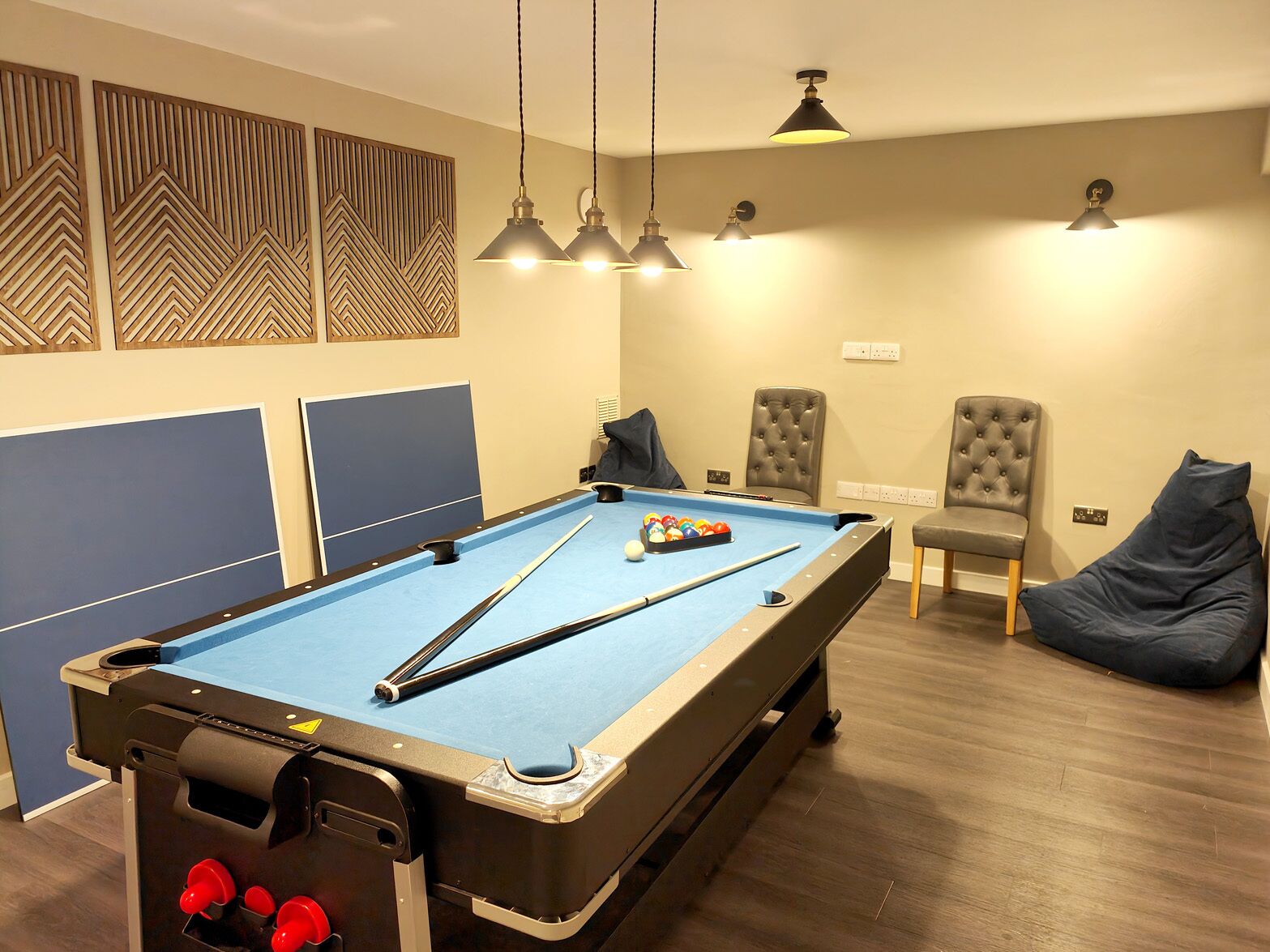 A pool table with a blue felt surface, two chairs, and a bean bag chair in a room with wooden flooring and wall decor.