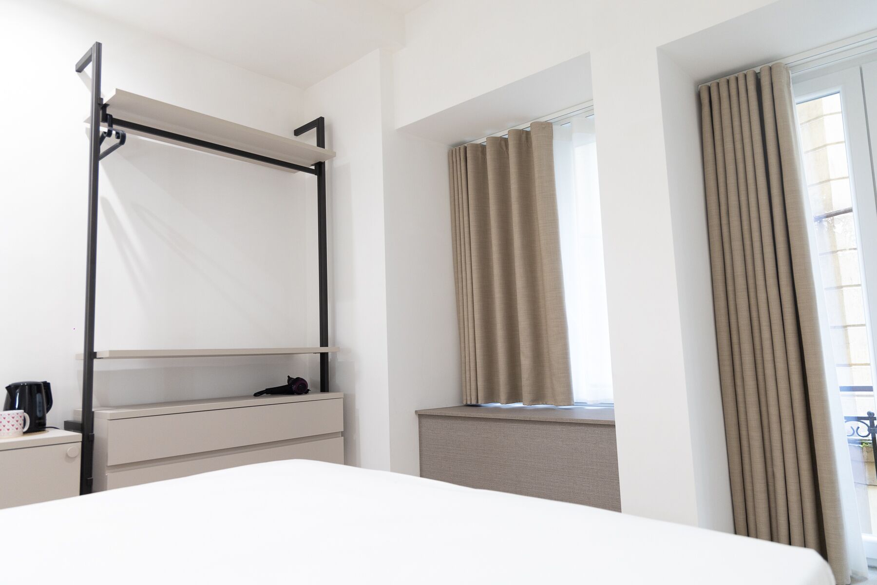 A hotel room with a bed, a desk with a coffee maker, a window with curtains, and a view of the outside.