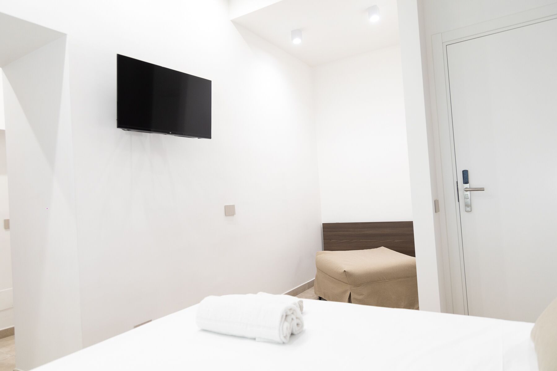 A modern hotel room with a bed, a flat-screen TV mounted on the wall, a wooden headboard, and a door with a digital lock.