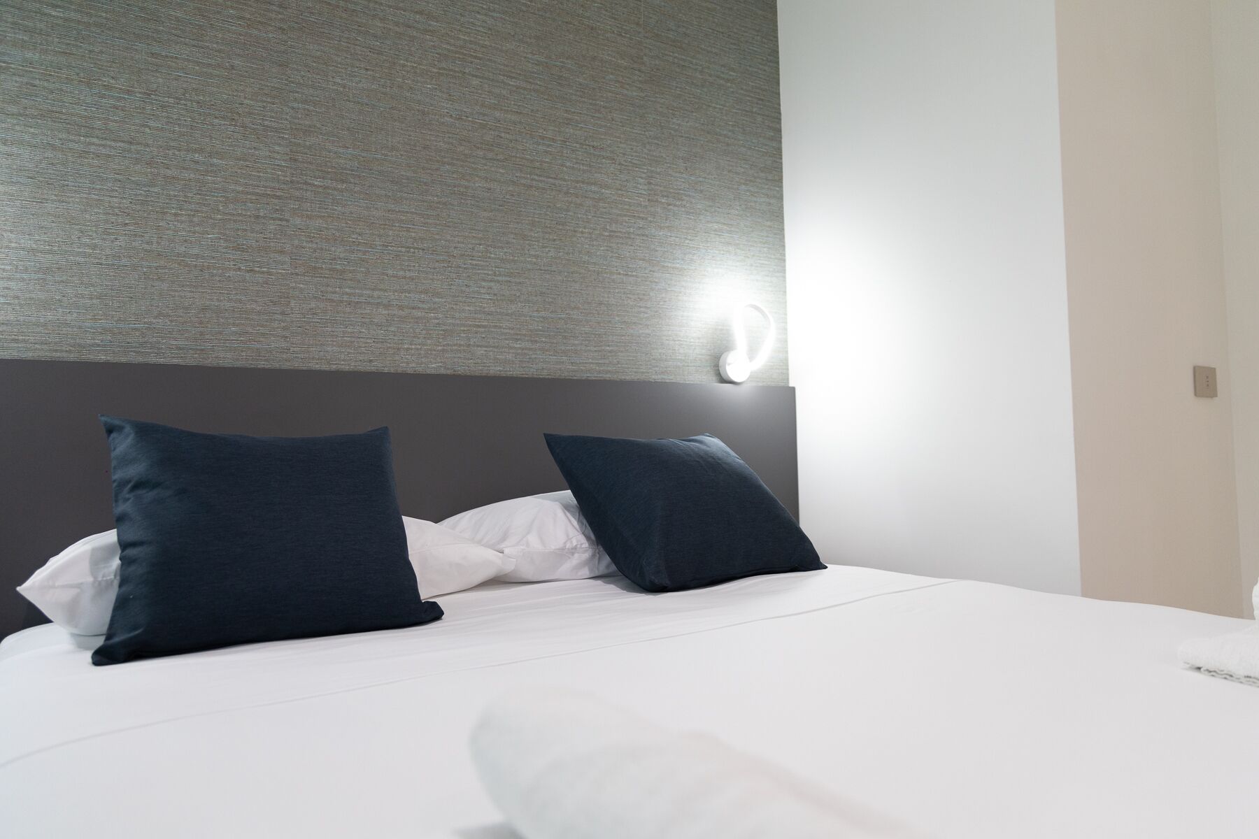 A neatly made bed with white bedding and two dark pillows, a textured wall above, and a light switch on the wall.