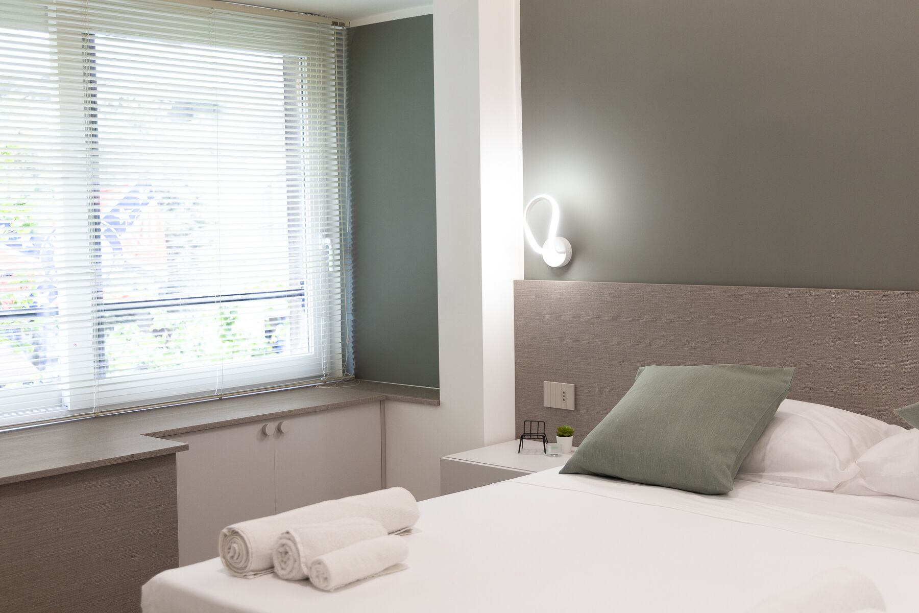 A hotel room with a bed, a nightstand, a window with blinds, and a wall-mounted light.
