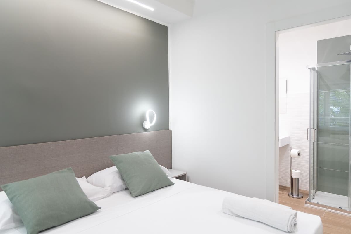 Una habitación de hotel moderna con una cama, almohadas verdes, una mesita de noche, una lámpara de pared y una zona de ducha de vidrio.