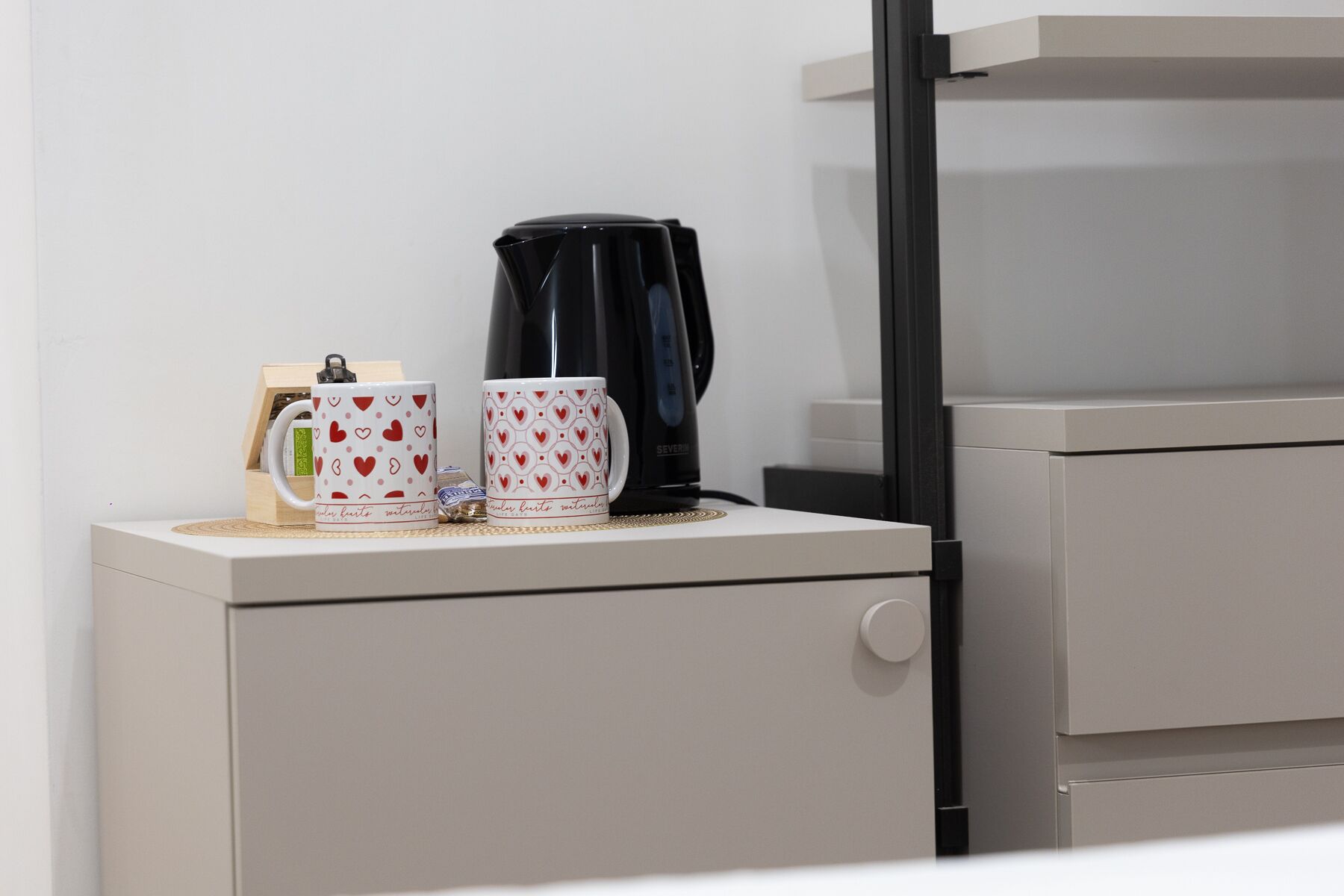 A small kitchenette with a coffee maker, two mugs, and a small wooden box on a white cabinet.