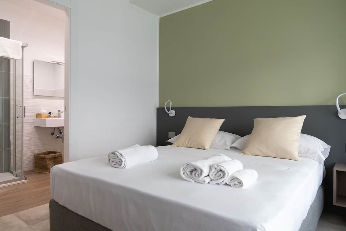 Habitación de hotel con cama, dos almohadas, una mesita de noche, una lámpara de pared, baño con lavamanos y espejo, y zona de ducha.