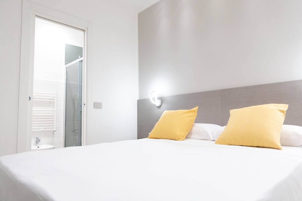 Habitación de hotel con una cama, dos almohadas, una mesita de noche y una lámpara fijada en la pared.