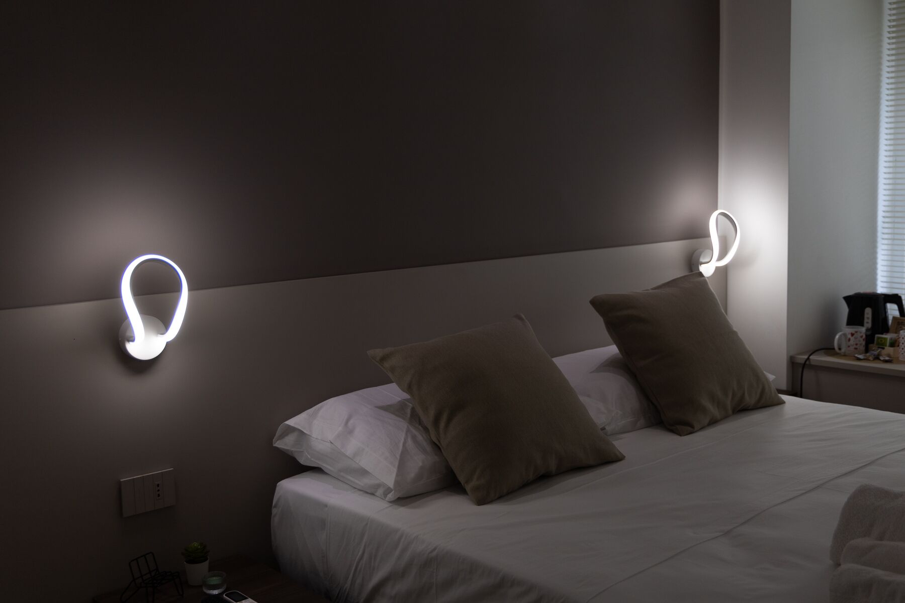 A modern hotel room with a bed, two pillows, a bedside table with a lamp, and a wall-mounted light fixture.