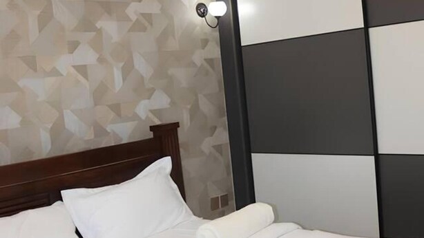 Un lit bien fait, avec du linge de lit blanc et des oreillers, une tête de lit en bois et un mur orné d’un motif géométrique, ainsi qu’un luminaire.