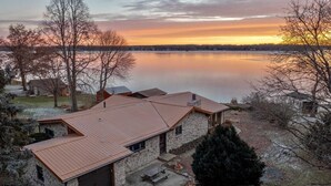A stone house with a metal roof, a garage, and a view of a calm lake at sunset. - West World (Beaver Dam)