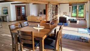 A spacious living area with a wooden dining table, comfortable seating, a stone wall, and large windows offering a view of the outdoors. - West World (Beaver Dam)