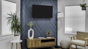 Ein modernes Wohnzimmer mit Flachbildfernseher an einer Wand mit geometrischem Muster, einem Holzkonsolentisch mit Dekorationsgegenständen, einem Rattan-Sessel und einer Topfpflanze.