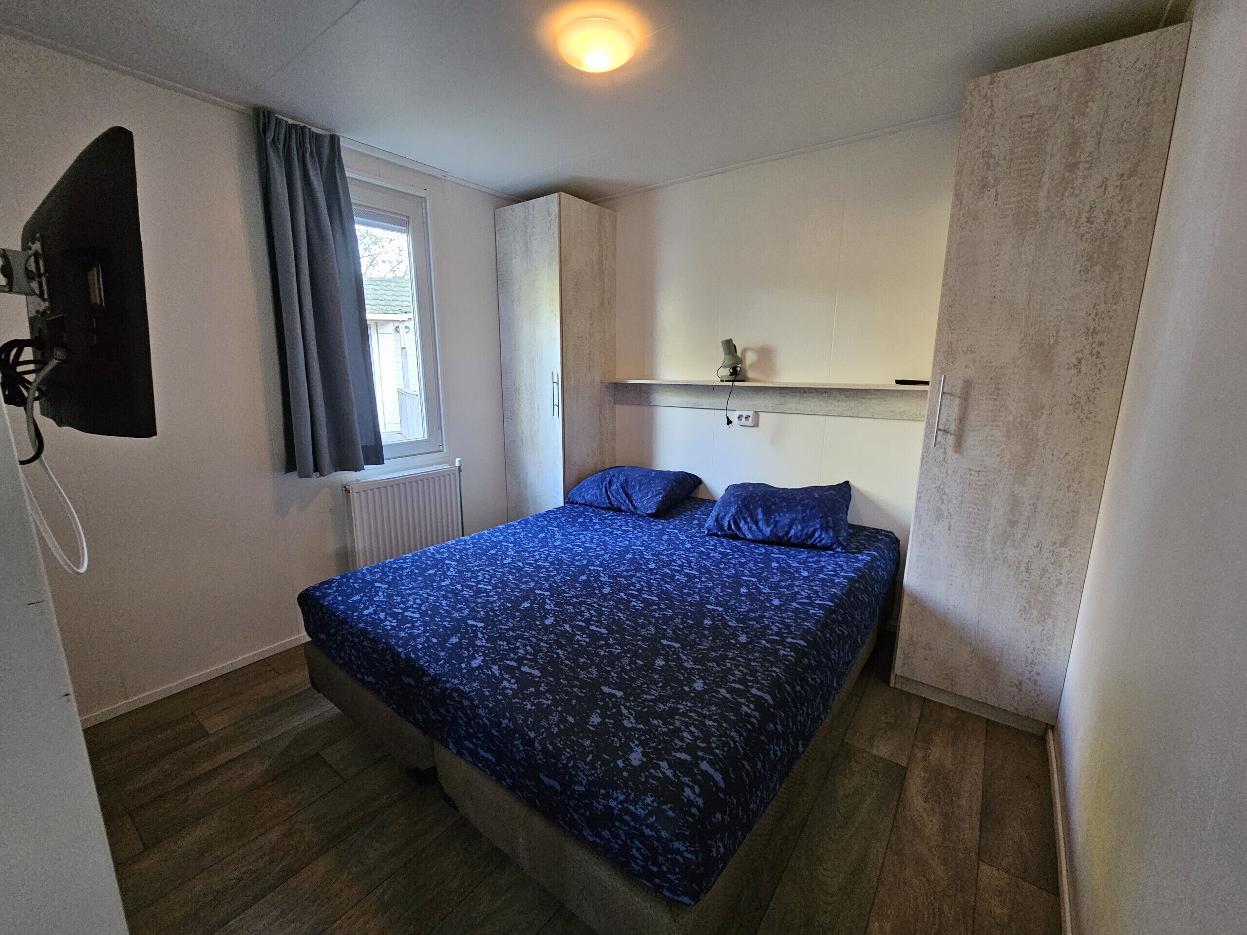 Une chambre à coucher avec un lit, une fenêtre avec des rideaux, une armoire et un téléviseur fixé au mur.