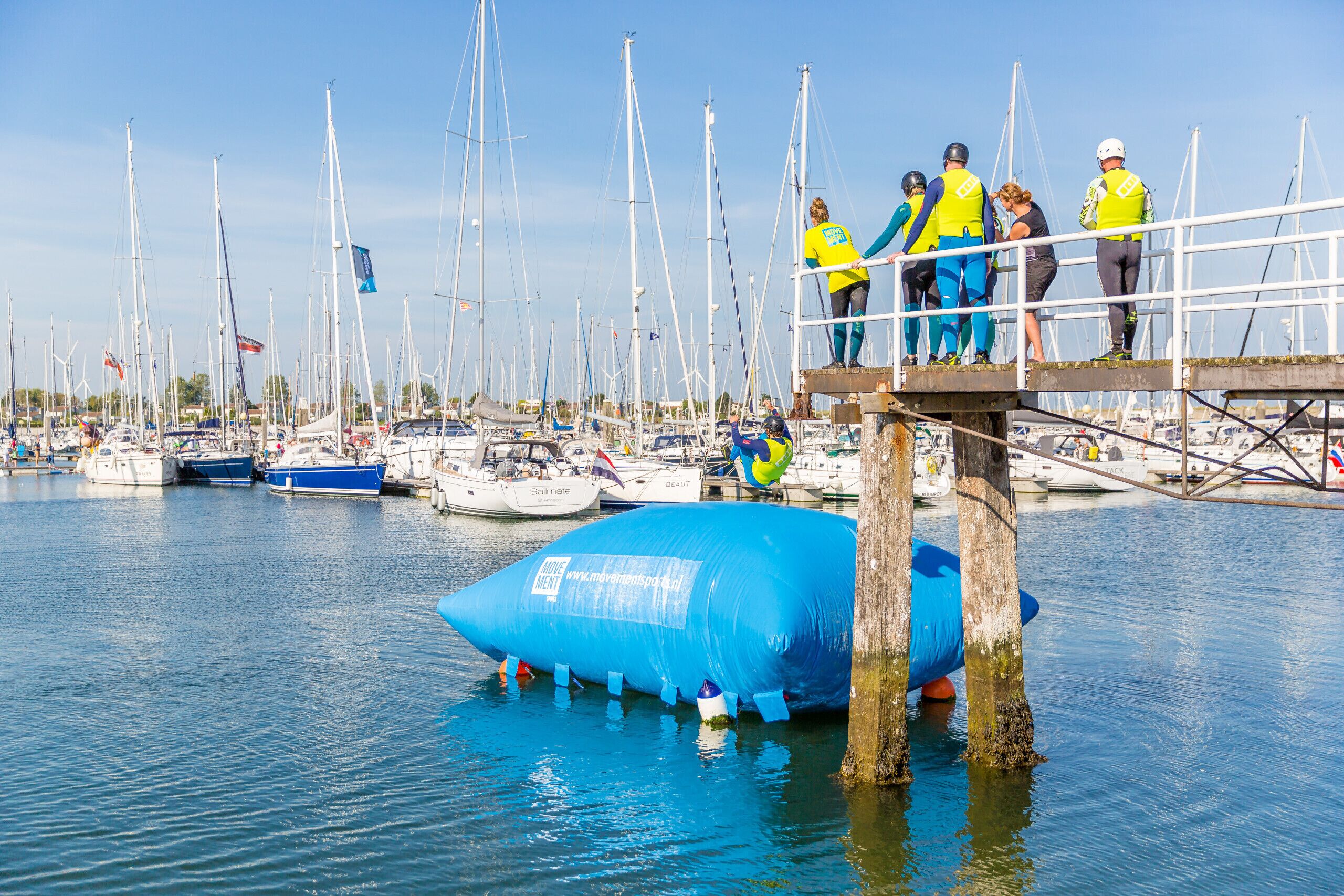 A marina with multiple sailboats docked, a large blue inflatable structure, and people wearing safety gear on a platform.