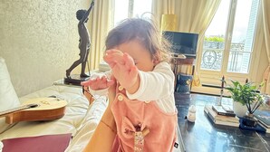 Un enfant est assis sur un canapé dans une pièce où se trouvent une statue, une guitare et une fenêtre avec des rideaux.