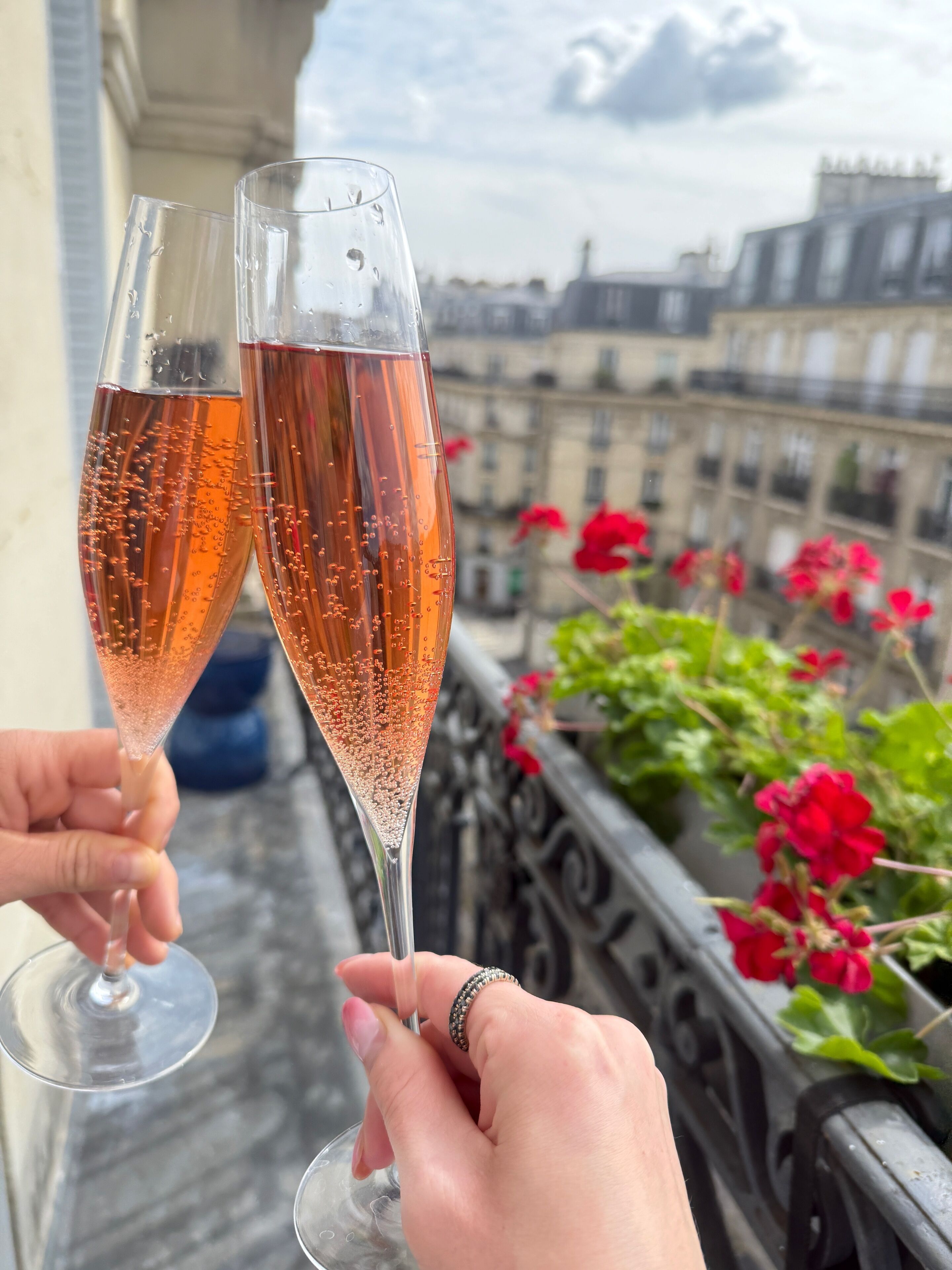 Two champagne flutes filled with a bubbly beverage, held by hands, with a balcony railing and red flowers in the background.