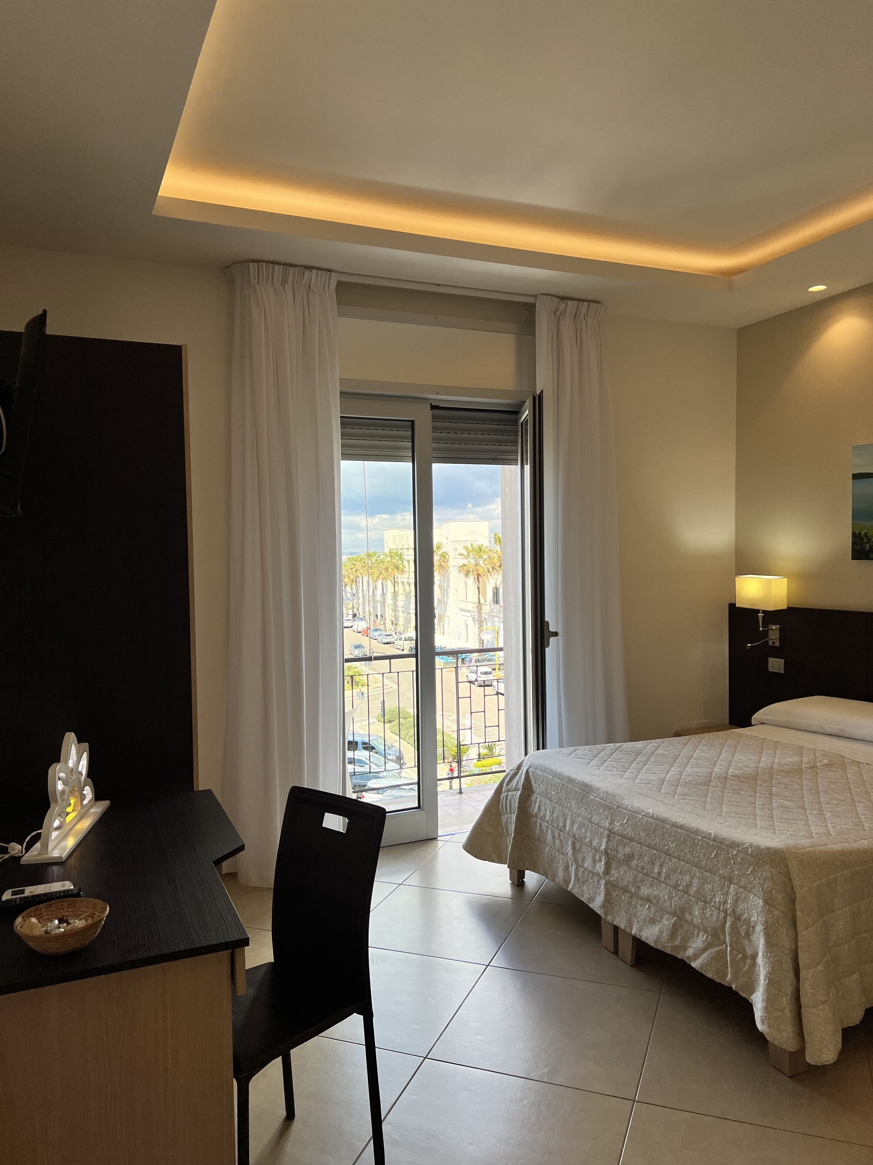 A hotel room with a bed, a desk, a chair, and a balcony with a view of palm trees and buildings.