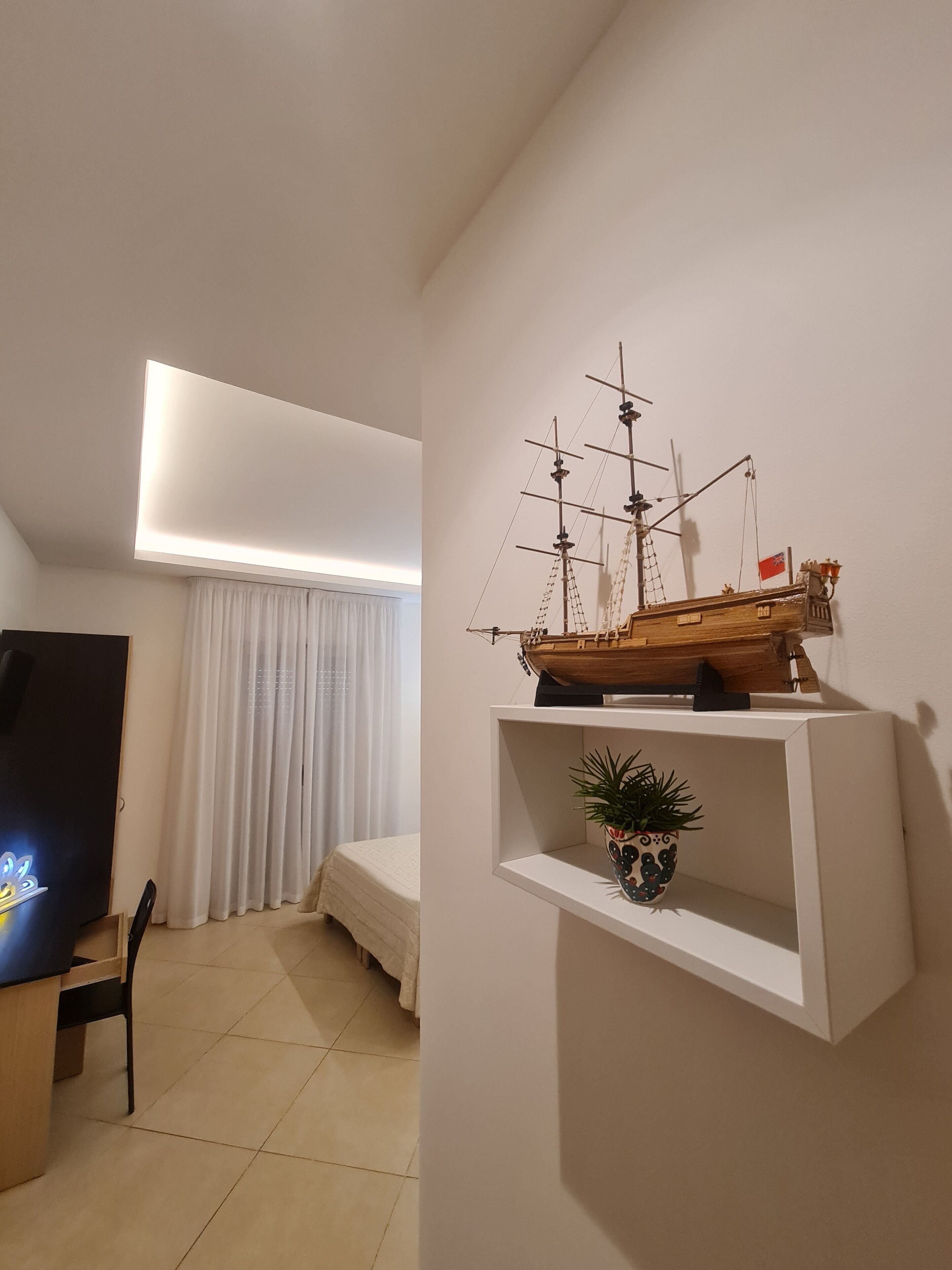 A bedroom with a bed, a desk, a chair, a ship model, and a shelf with a plant.