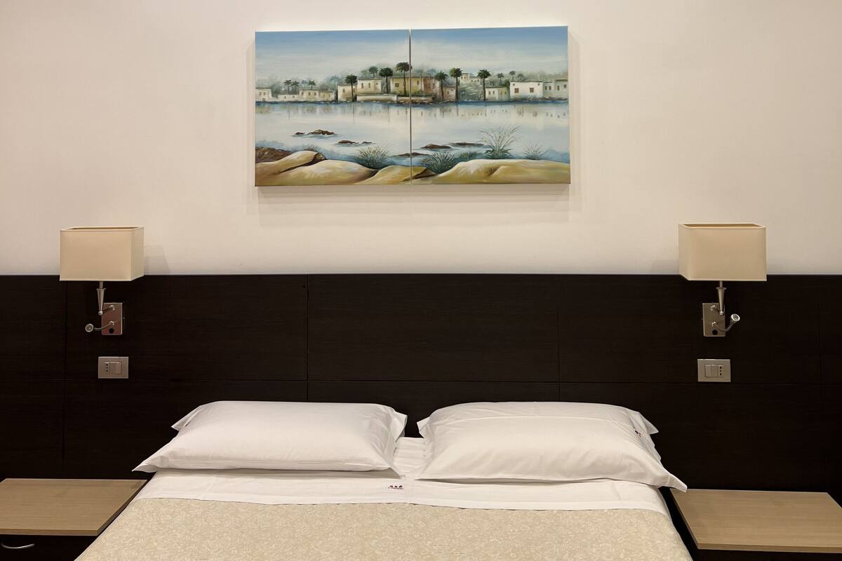 A hotel room with a bed, two bedside tables, a painting on the wall, and recessed ceiling lights.