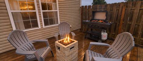 Un patio exterior con sillas Adirondack, un hogar para fogata cuadrado y una parrilla.