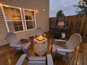 An outdoor patio with Adirondack chairs, a square fire pit, and a barbecue grill.