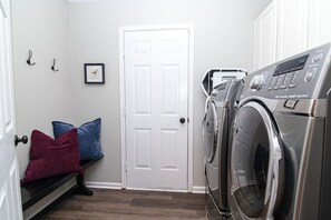 A laundry room with a front-loading washer and dryer, a bench with pillows, a white door, and wall-mounted hooks.