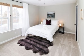 A bedroom with a bed, a window with curtains, a nightstand with a lamp, and a painting on the wall.