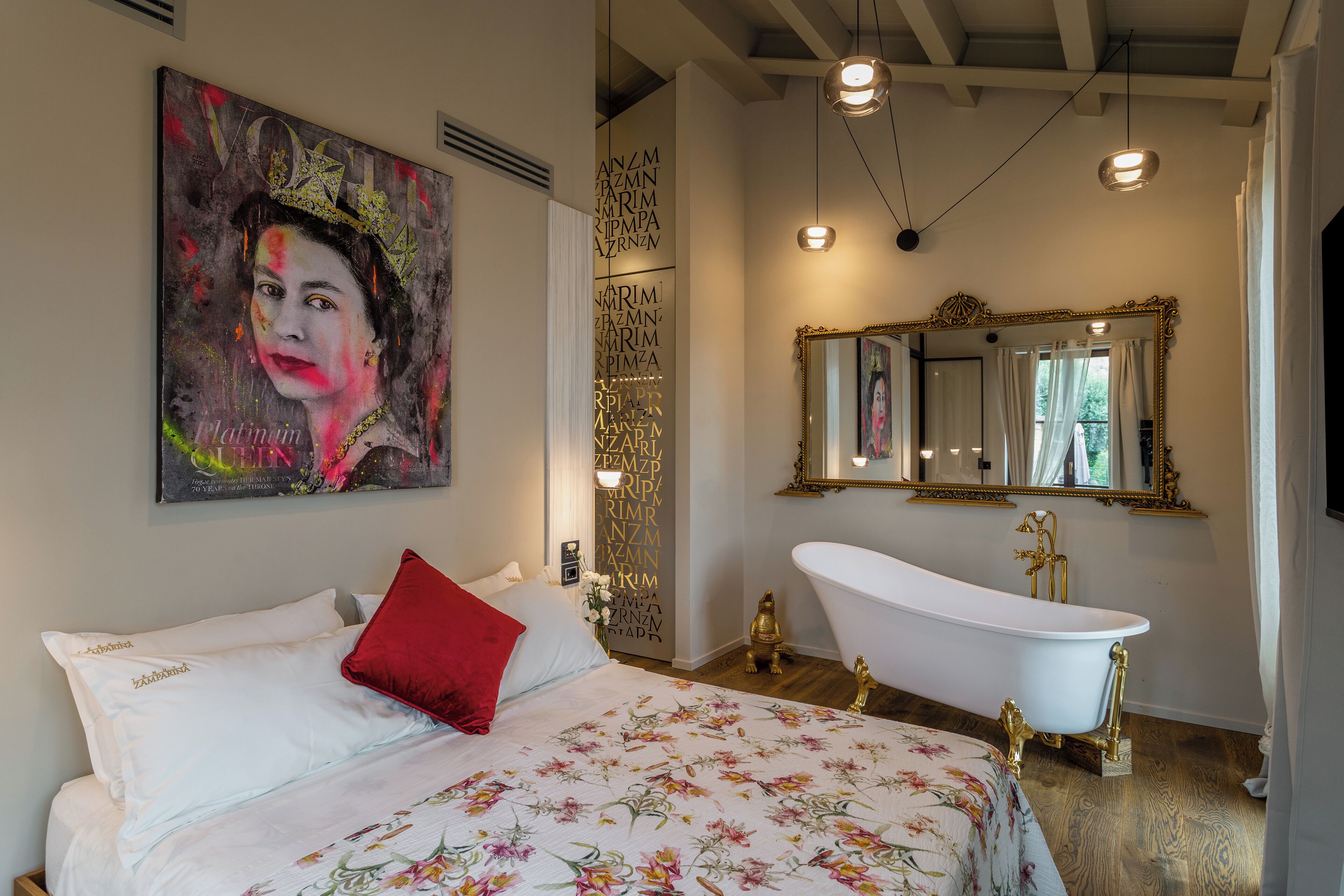 A bedroom with a large bed, a red pillow, a painting, a white bathtub, and a mirror.
