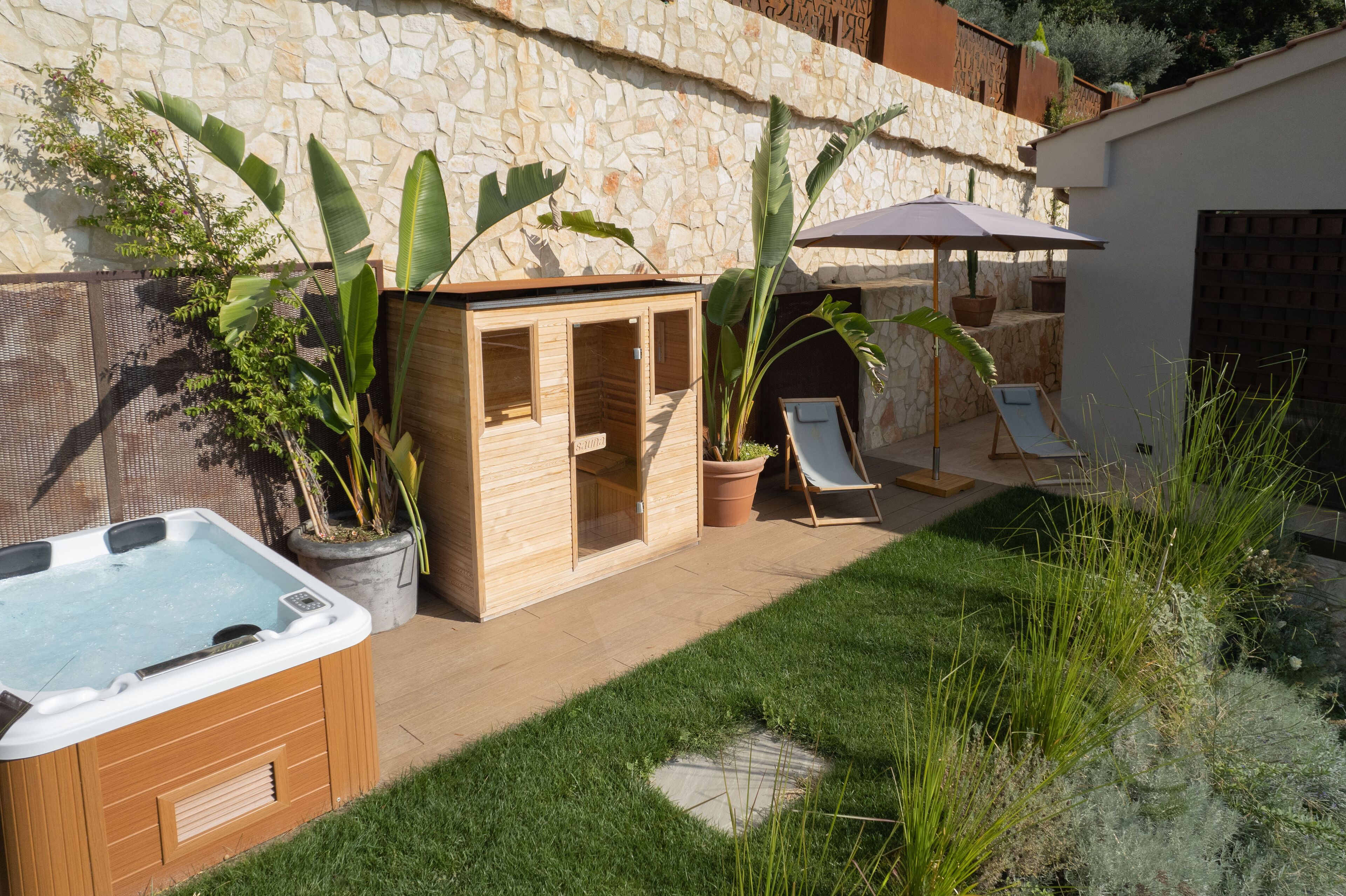 A hot tub, wooden sauna, and lounge chairs in a backyard with stone walls and potted plants.