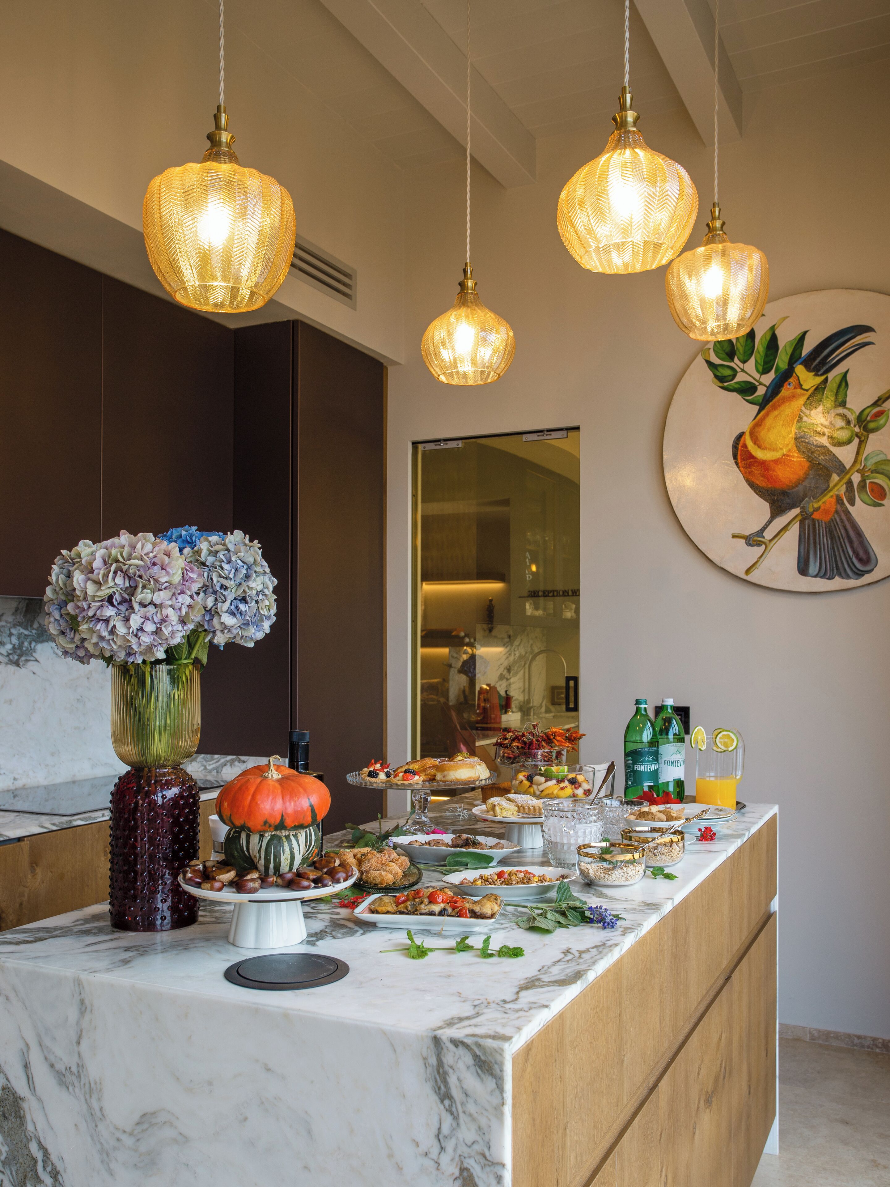 Un'area cucina ben illuminata con un piano di lavoro in marmo, diversi alimenti e elementi decorativi come un vaso di fiori e un dipinto di un uccello.