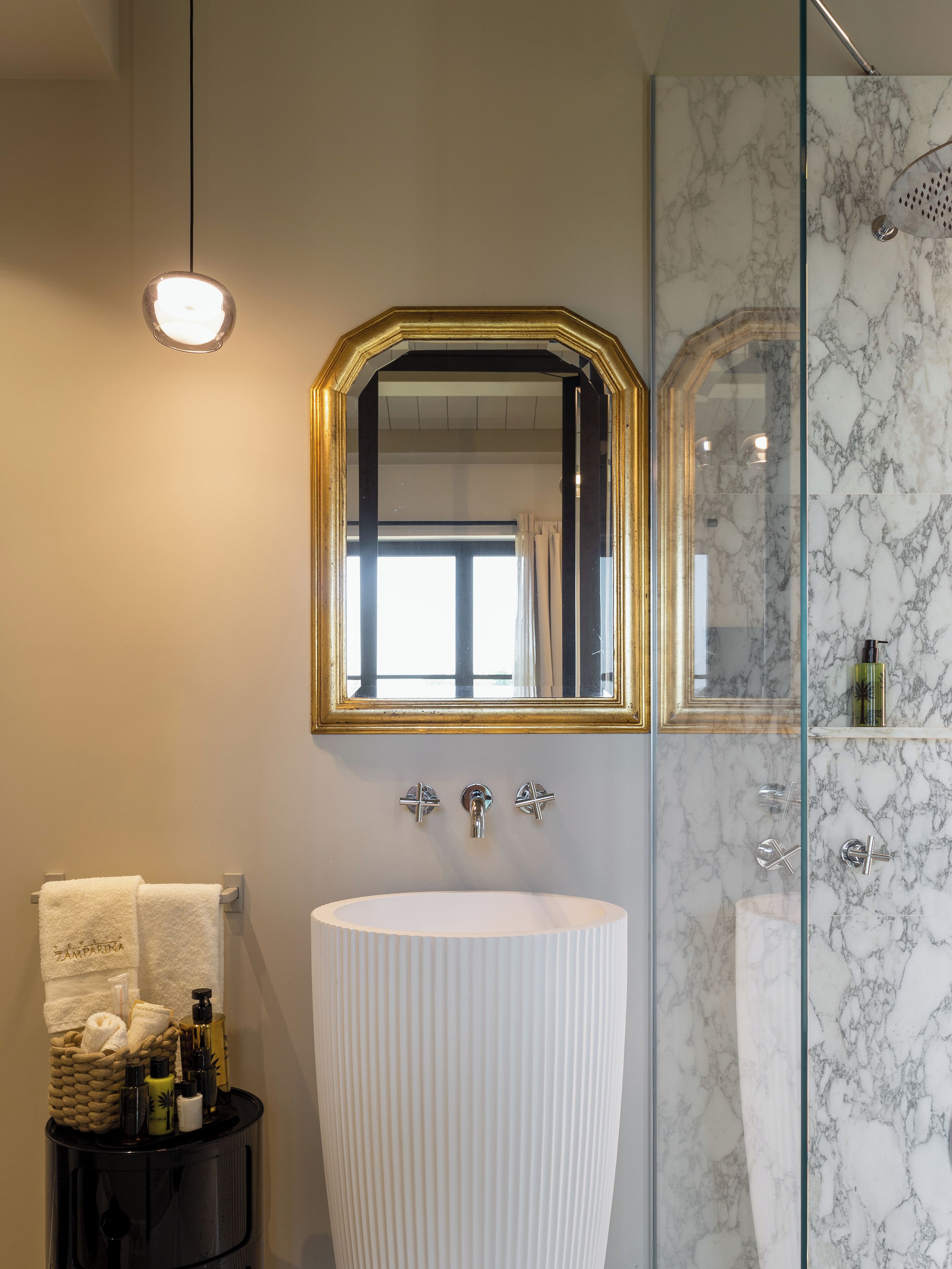 A bathroom with a large oval sink, a mirror with a gold frame, and a shower with marble walls.