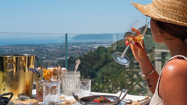 A person seated at a table with a view of a cityscape, a glass of drink, and a pan with food. - Tenuta Zamparina (Montignoso)