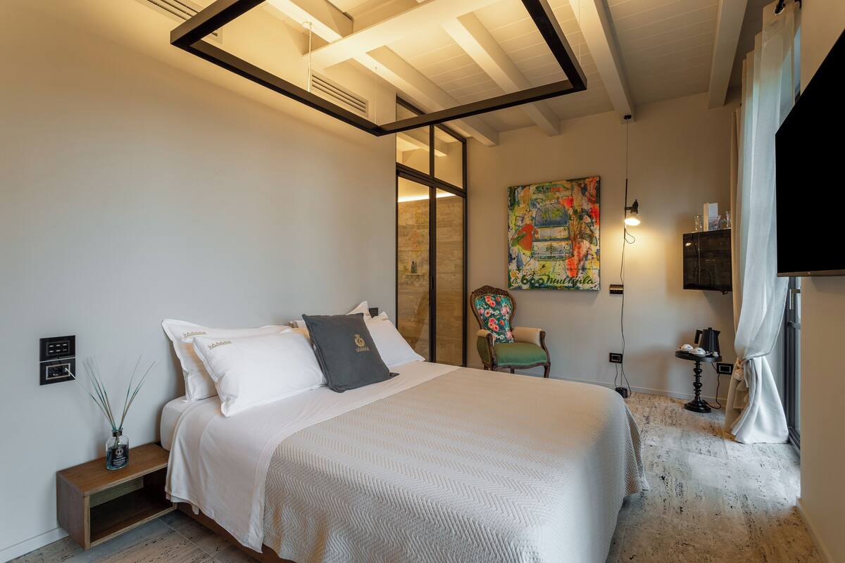 A bedroom with a large bed, a nightstand, a chair, a painting, and a TV.