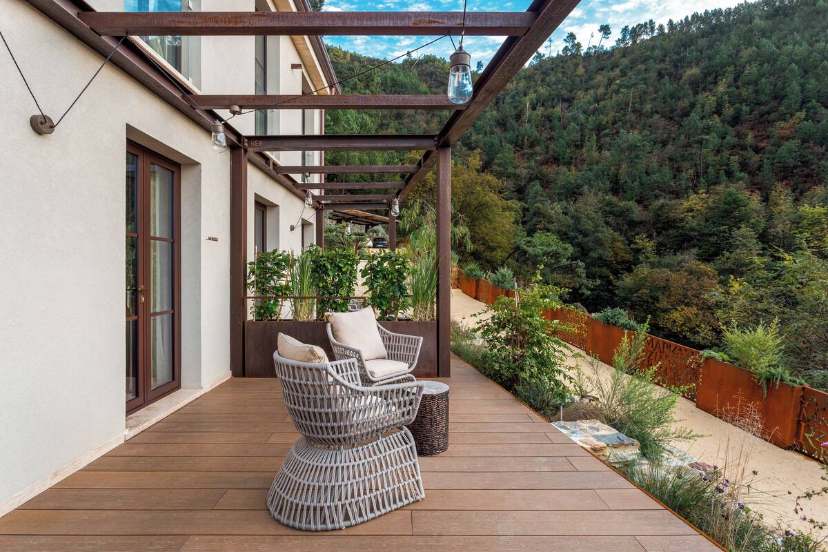 A covered outdoor seating area with wicker furniture, a wooden deck, and a view of a forested hillside.