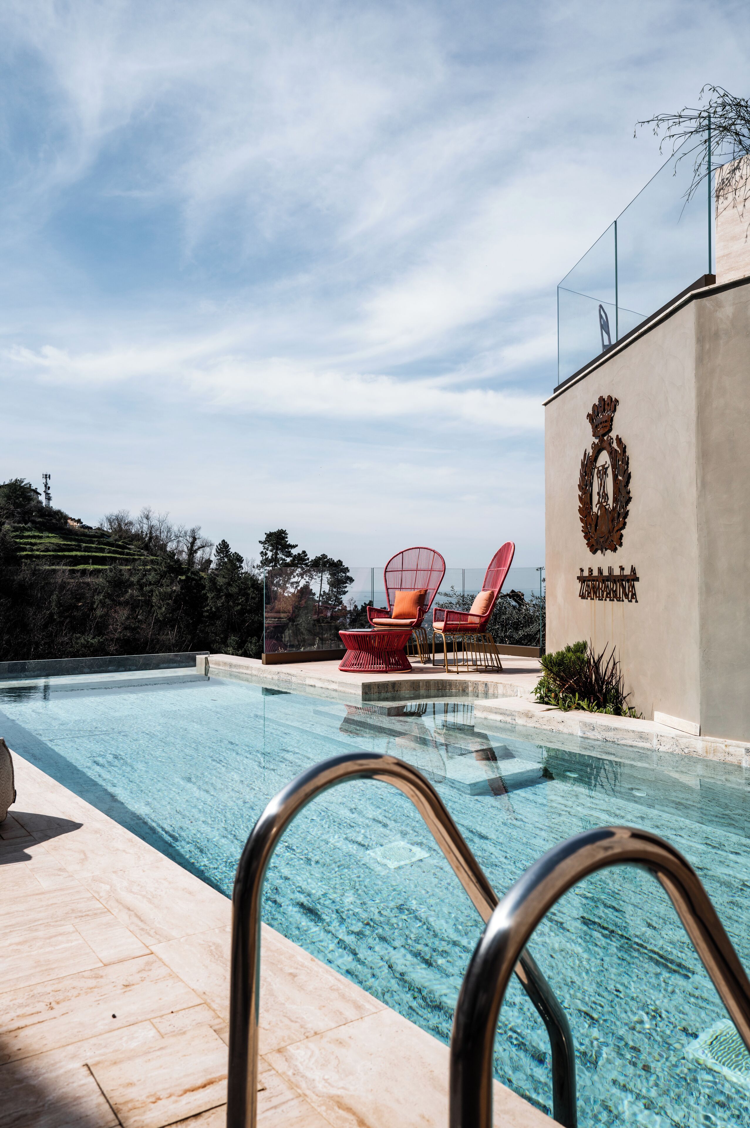 A swimming pool with clear water, surrounded by a tiled deck. There are two red chairs with a glass top near the pool. A building with a crest emblem is visible in the background.