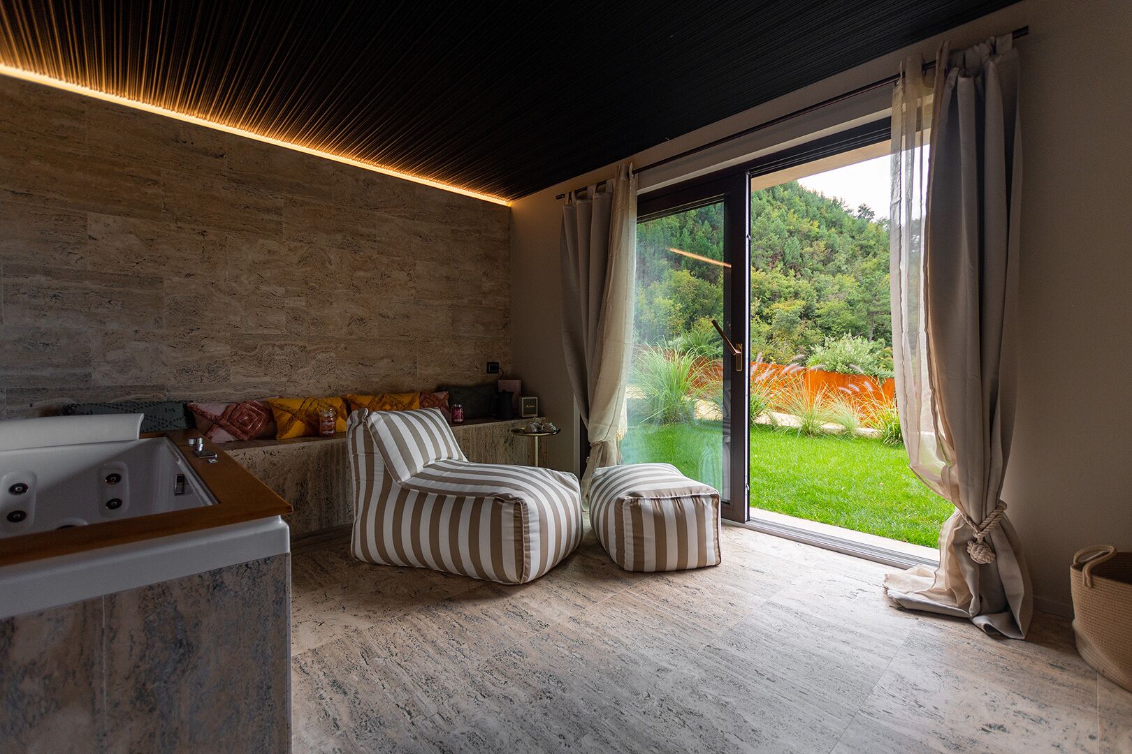 Un bagno moderno con una parete in pietra, una vasca da bagno, cuscini a righe e vista su un giardino verde attraverso ampie vetrate.