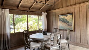 Una habitación con paneles de madera, una mesa redonda y cuatro sillas, una araña, un ventanal con cortinas y un cuadro de paisaje en la pared.