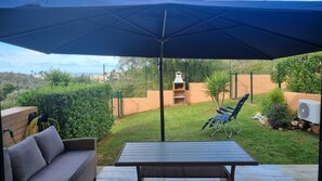 Eine Terrasse im Freien mit Tisch und Stühlen, einem großen Sonnenschirm und Blick auf einen Garten und Hügel im Hintergrund.