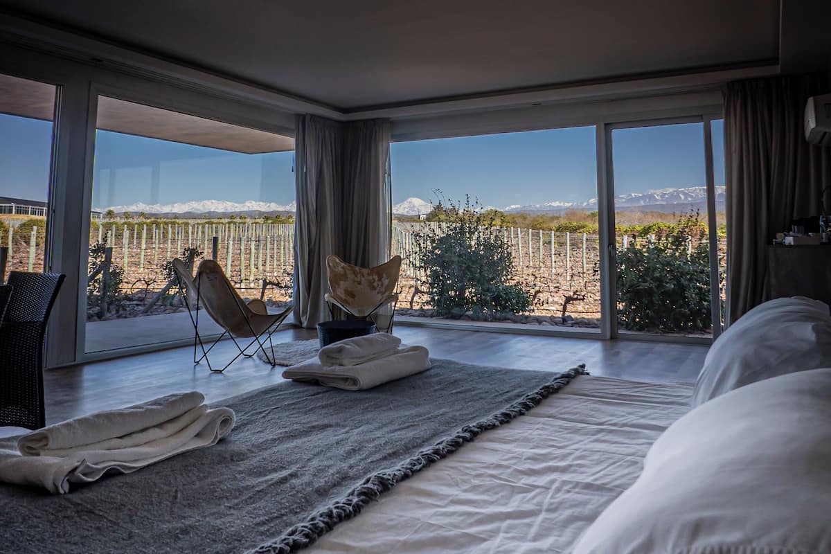 Quarto com uma cama grande, uma cadeira, um tapete e vista para uma paisagem montanhosa através de grandes janelas.