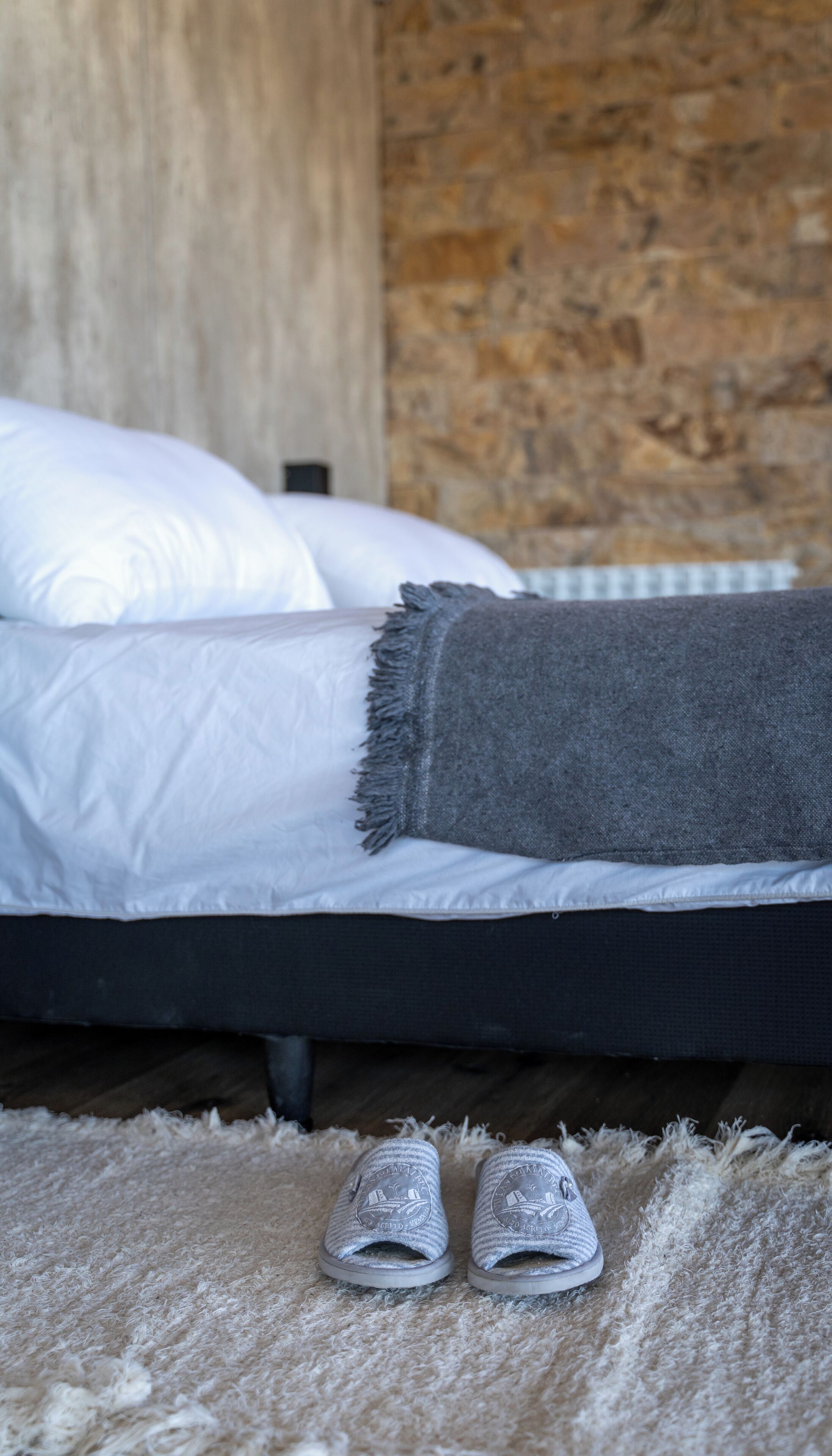 A bed with white pillows and a gray blanket, a pair of slippers on a fluffy rug, and a wooden wall in the background.