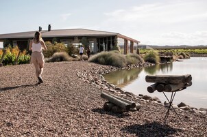 Una persona caminando por un sendero de grava cerca de un lago, con una casa al fondo y leña apilada junto al agua.