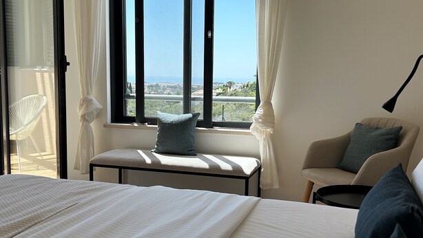 Ein Hotelzimmer mit einem großen Bett, einem Fenster mit Meerblick, einem Sessel und einem kleinen Tisch.