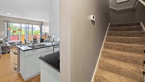 A modern kitchen with white cabinets, a black countertop, and a stainless steel sink. There is a staircase with carpeted steps leading to a landing.