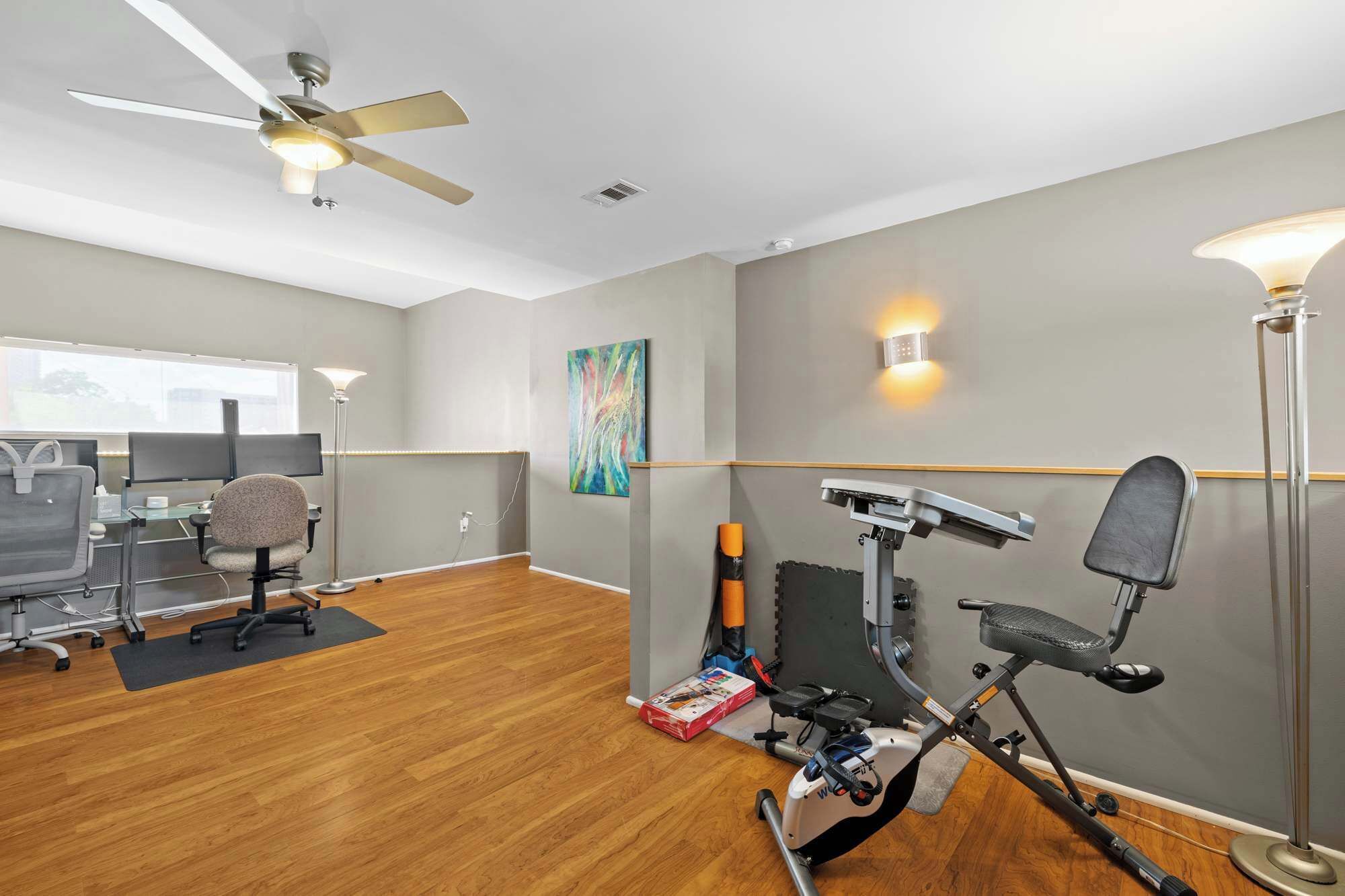 A spacious room with a ceiling fan, a treadmill, exercise equipment, a desk with a computer, and a chair.